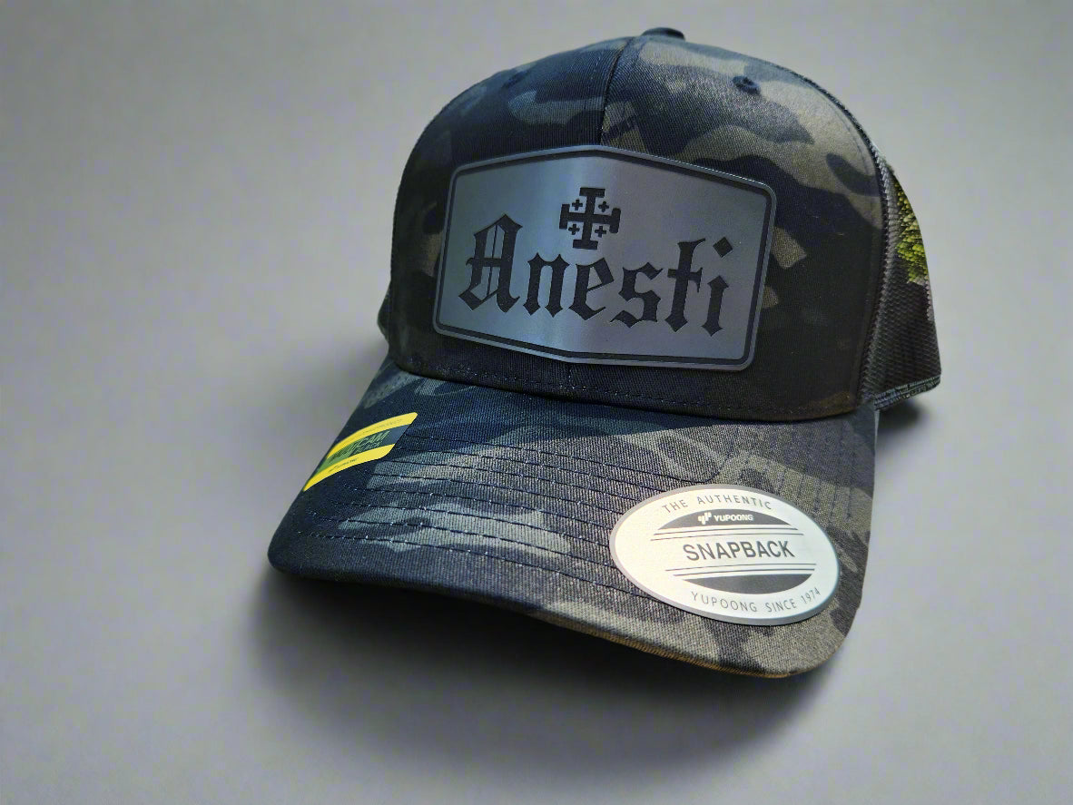 Black and gray camouflage cap with 'Anesti' logo on a wooden surface with a natural background