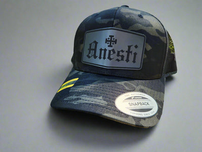 Black and gray camouflage cap with 'Anesti' logo on a wooden surface with a natural background